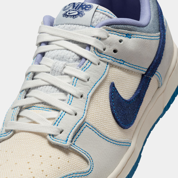 Nike Dunk Low Retro SE sneakers with a white and beige mesh upper, blue suede Swoosh, contrasting blue stitching, and white laces, close-up angled view.