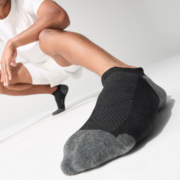 Feetures Elite Max Cushion No Show Tab socks in black and gray with targeted compression and seamless toe, worn by athlete during stretch.