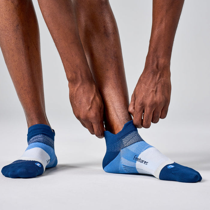 Feetures Elite Ultra Light No Show Tab socks in blue and white, worn by person adjusting, highlighting seamless toe design.