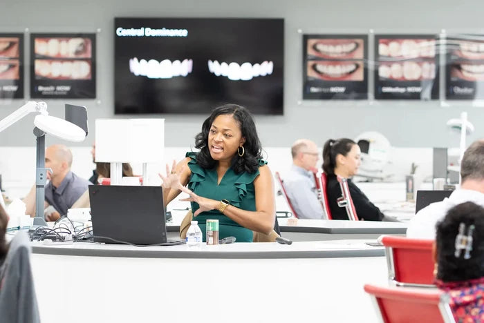 Woman speaking at a desk in a classroom setting with educational posters on the wall.