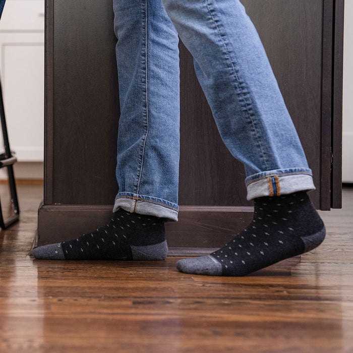 Feetures Everyday Men's Max Cushion Crew socks in black with gray accents and subtle polka dot pattern, worn casually indoors.