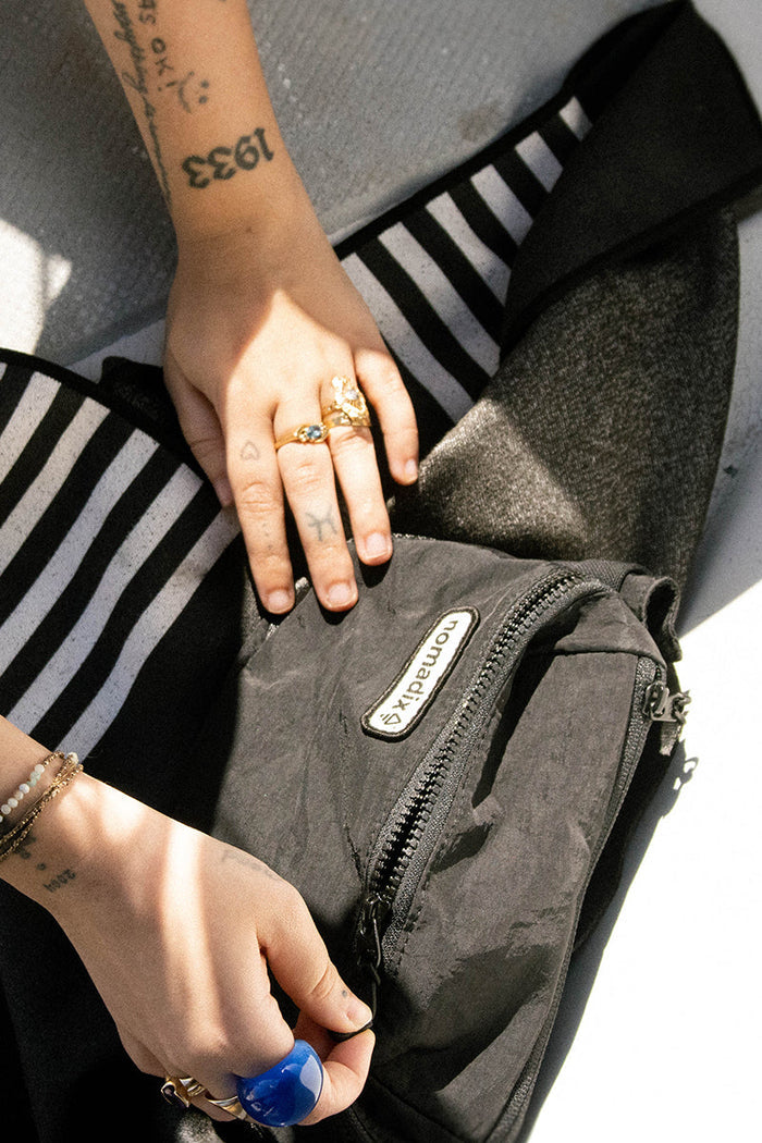 Nomadix Fanny Pack in black with zippered compartments and branded logo, lifestyle context showing hands interacting with the pack.