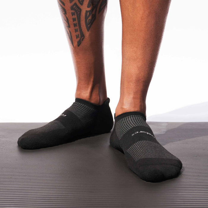 Feetures High Performance Max Cushion No Show Tab socks in black, featuring a cushioned design and mesh ventilation, worn by a person standing on a textured surface.