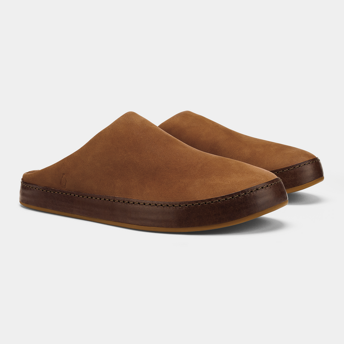 OluKai Ku'u slippers in warm brown suede with dark leather accents, side front angle showing stitch detailing and slip-on design.