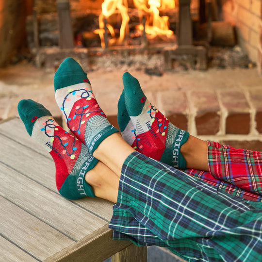 Feetures Limited Edition "Merry & Bright" Elite Light Cushion No Show Tab Socks in green, red, and festive light design, lifestyle context by a fireplace.