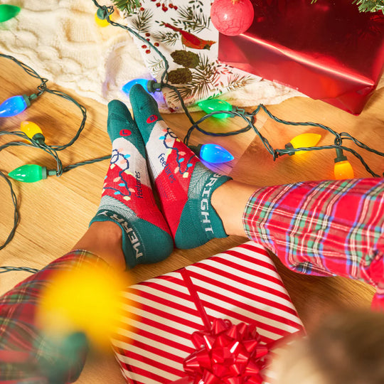 Feetures Limited Edition "Merry & Bright" Elite Light Cushion No Show Tab Socks, festive red, white, and green design with holiday graphics, worn by person sitting near Christmas lights and gifts.