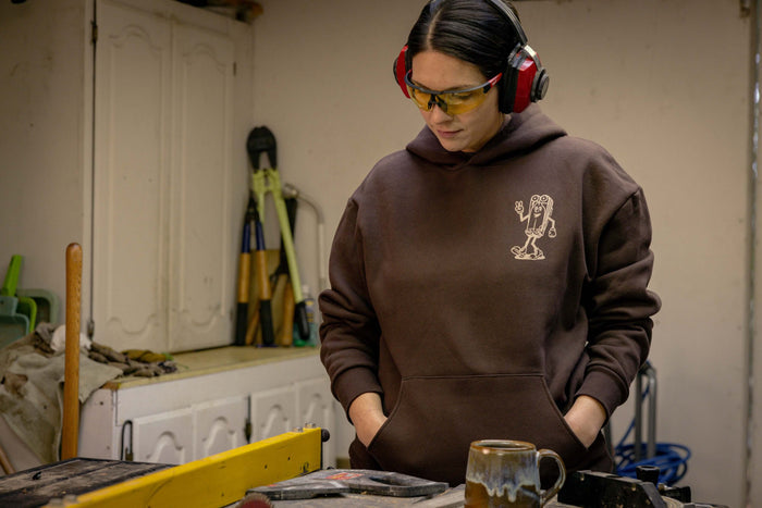 A person wearing a brown Leatherman Mr. Wave hoodie standing in a workshop with safety glasses and ear protection, looking down at a workbench with tools and a ceramic mug.