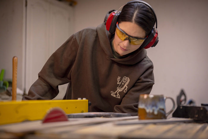 A person wearing a brown Leatherman Mr. Wave hoodie operating a table saw in a workshop, wearing safety glasses and earmuffs for protection.