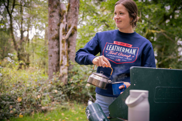 A person wearing a navy blue Leatherman PNW Crewneck sweatshirt pouring hot water from a kettle into a mug at a campsite surrounded by trees.