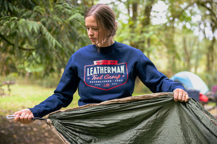 A person wearing a navy blue Leatherman PNW Crewneck sweatshirt setting up a hammock at a forest campsite with a tent in the background.